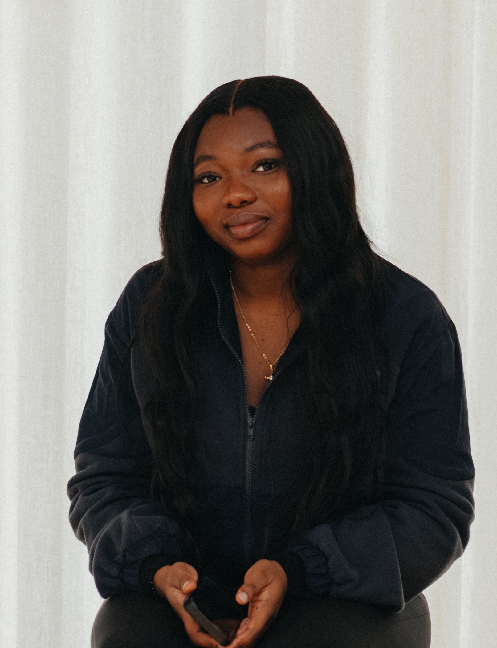 Portrait of a woman with long dark hair sitting indoors against a light background. Sarah Bakare