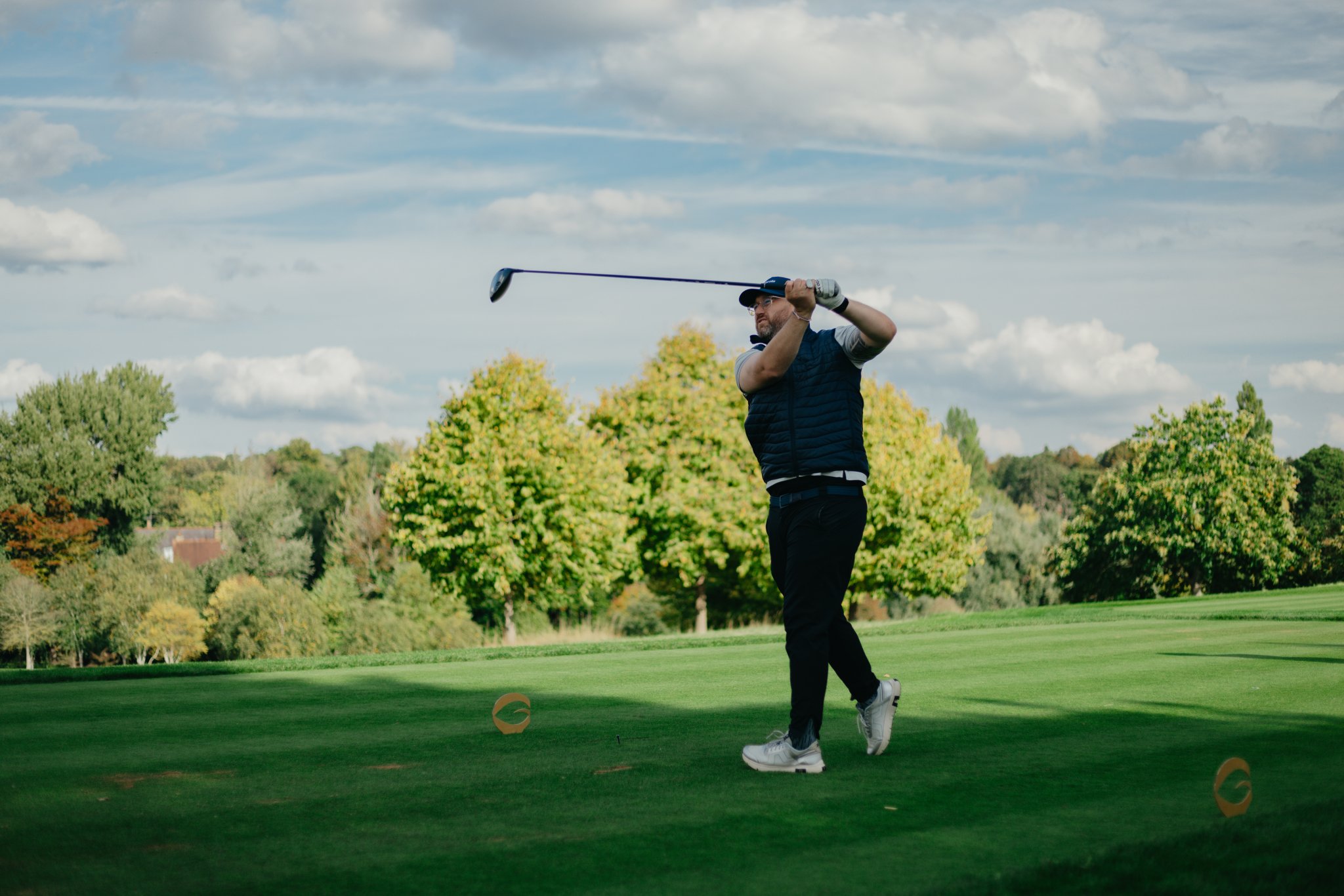 Golfer swinging a club on a sunny golf course with trees in the background.