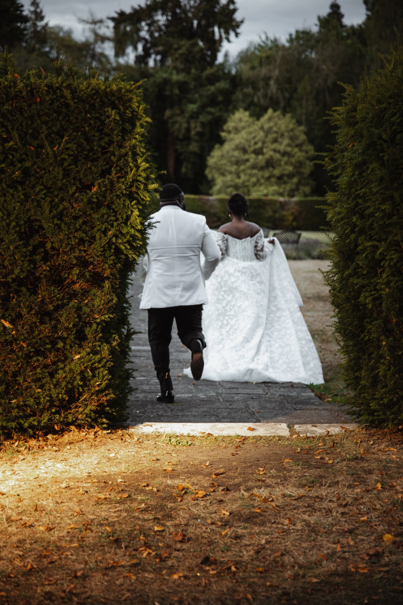 Bride and groom walking away together along a garden pathway surrounded by tall hedges.