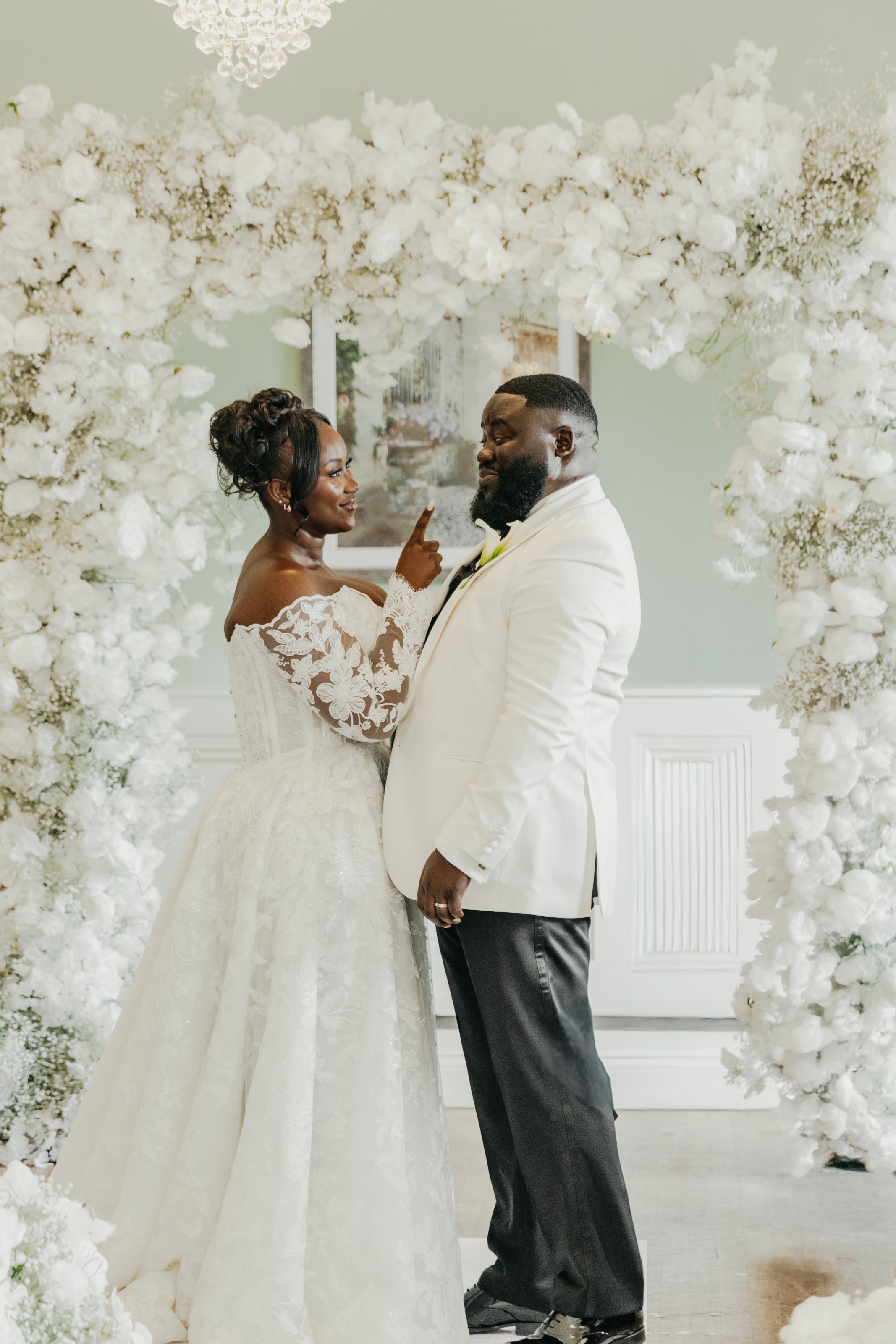 Bride and groom smiling at each other beneath an elaborate white floral wedding arch.