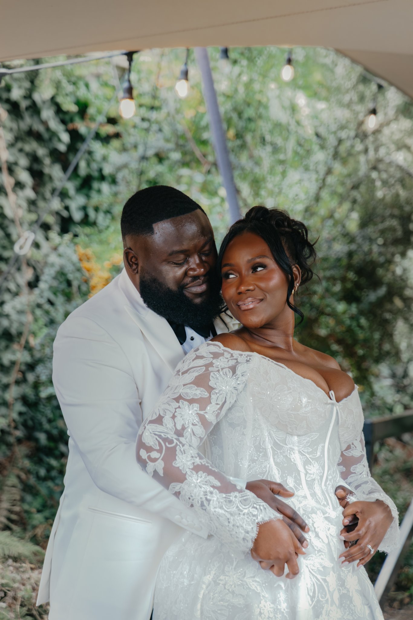 Couple embracing in wedding attire beneath trees and hanging lights.