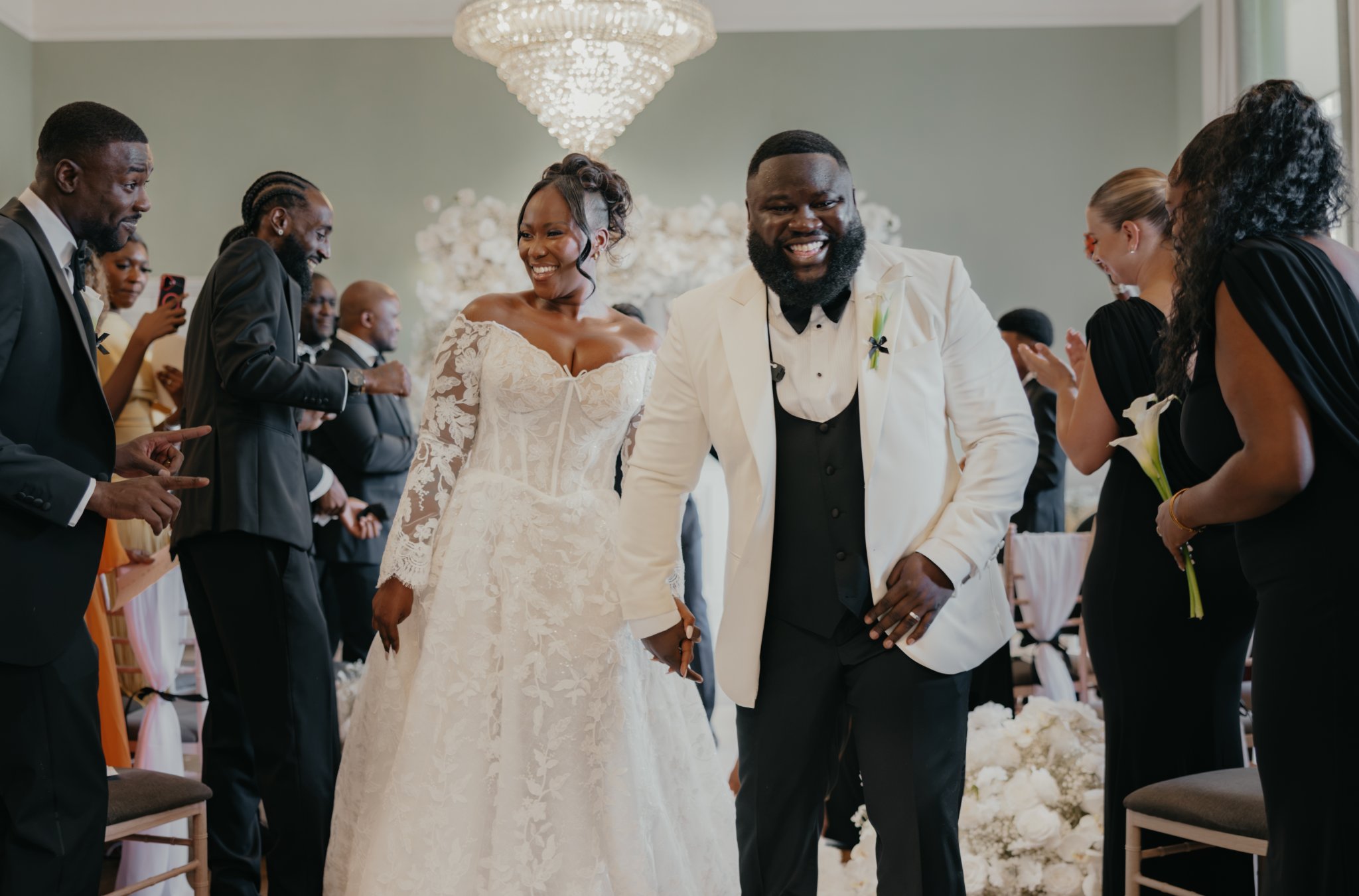 Bride and groom walking hand in hand through their wedding reception, smiling beneath an ornate chandelier.