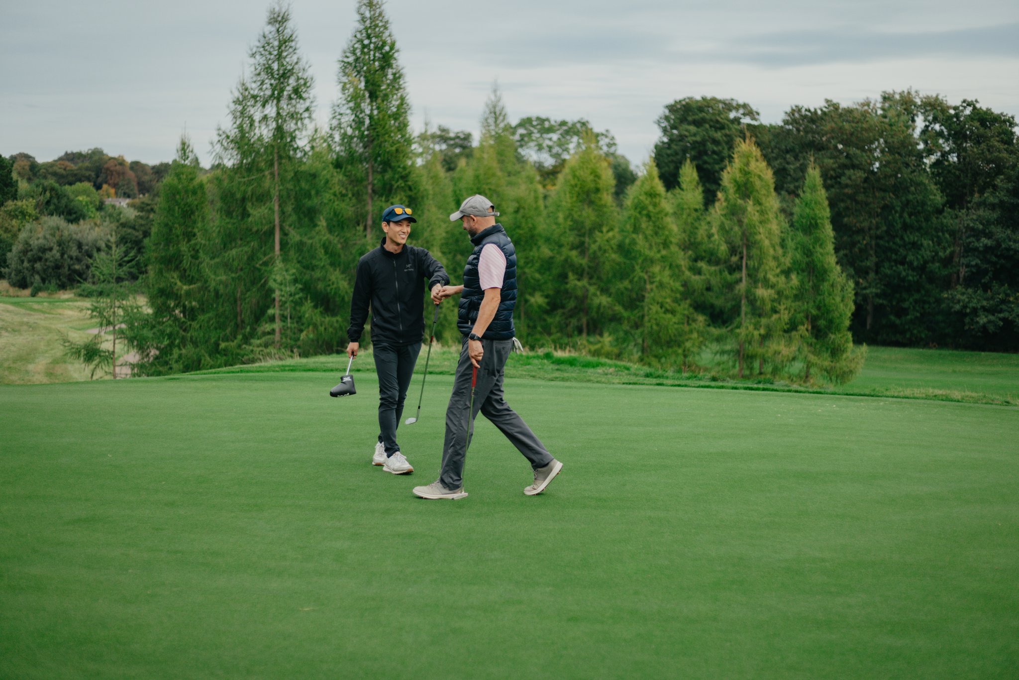 Two men walking together across a golf course surrounded by green trees.