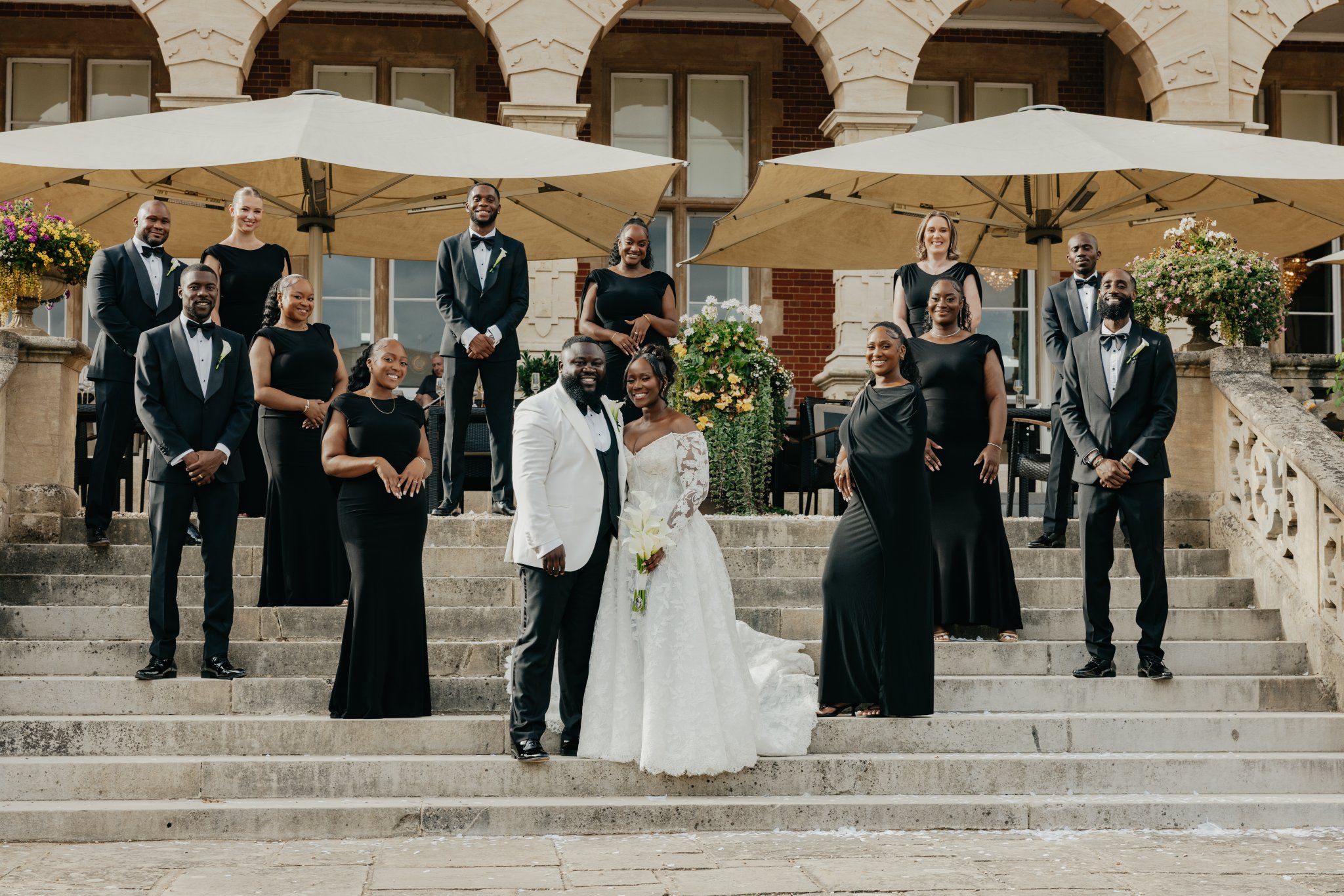 Bride and groom posing on outdoor steps surrounded by their wedding party dressed in black.