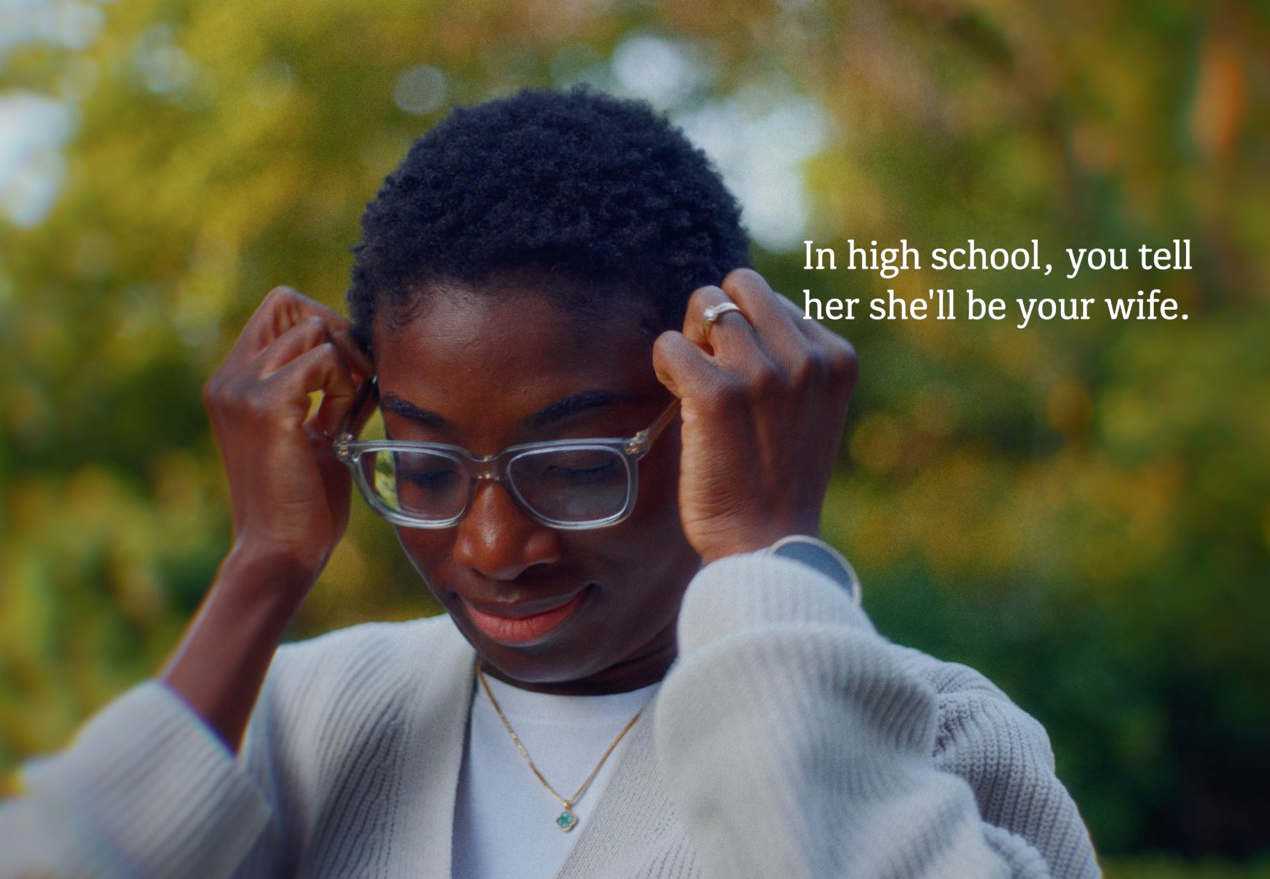 Close-up of a black woman with short natural hair adjusting her glasses with both hands, wearing a light cardigan against a softly blurred outdoor background.