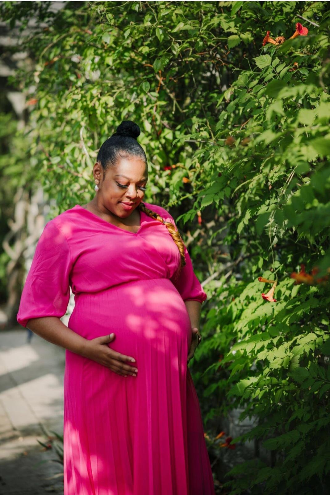 Pregnant woman in a bright pink dress standing in a garden and holding her belly.