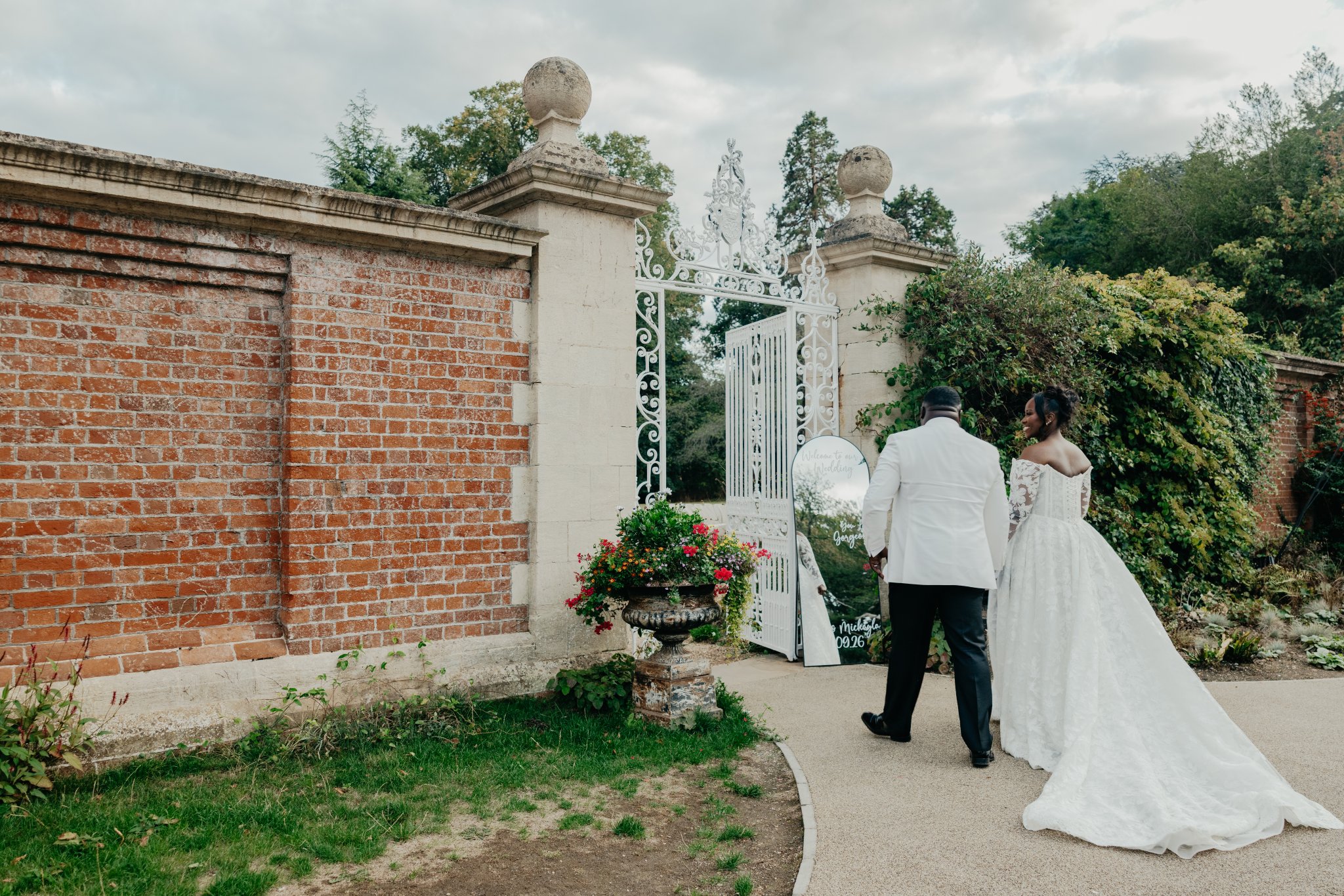 Groom walking towards an ornate garden entrance with brick pillars, white gates and floral decorations.