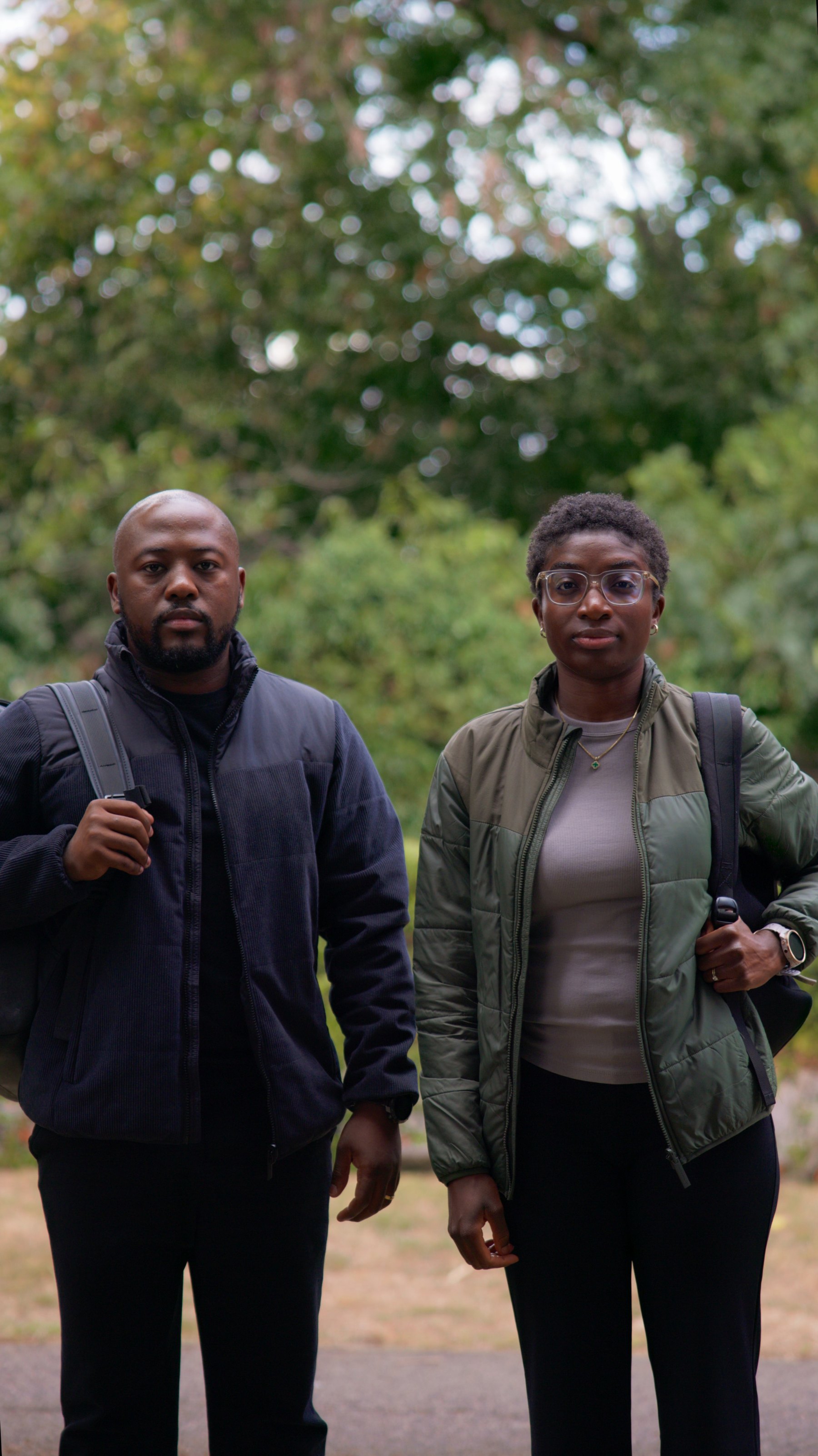 Two people standing together outdoors with soft green foliage behind them.